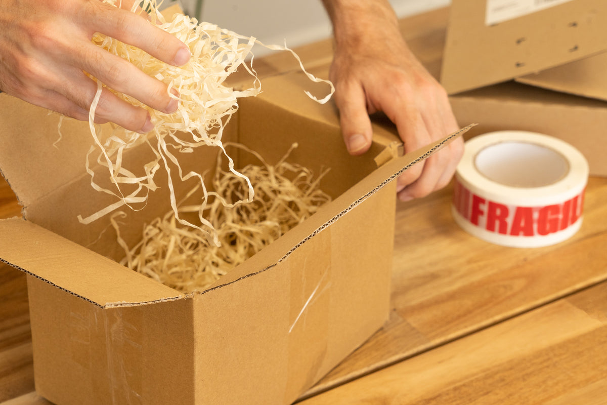 Hands packing shredded wood wool into an open cardboard shipping box, with a roll of red FRAGILE tape on the table beside it