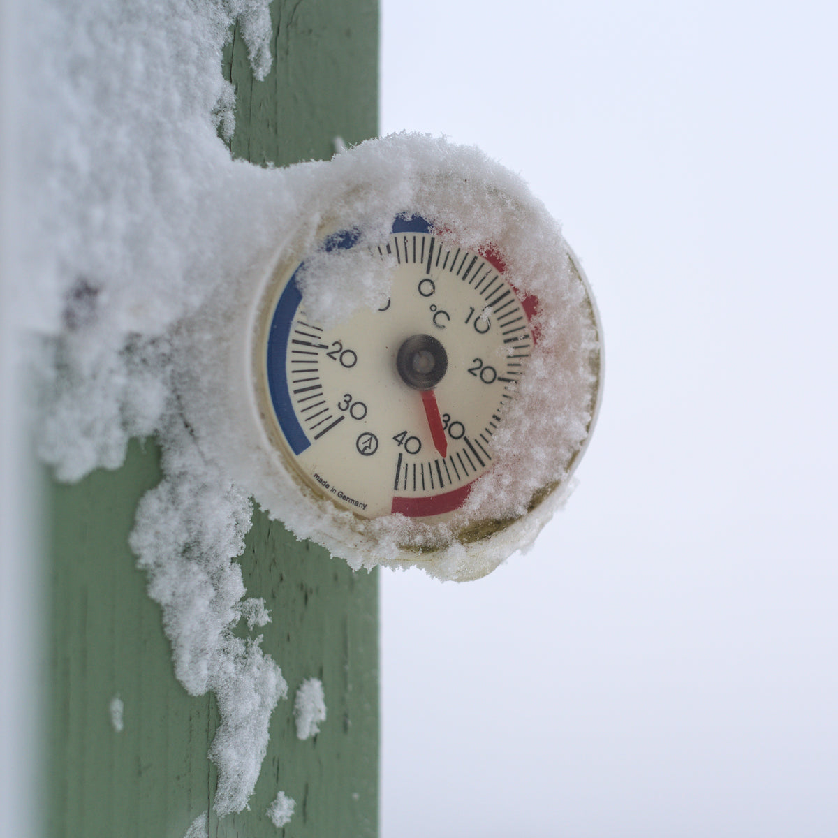 A round dial thermometer mounted on a green wooden window frame, half buried under fresh snow, against a plain white sky