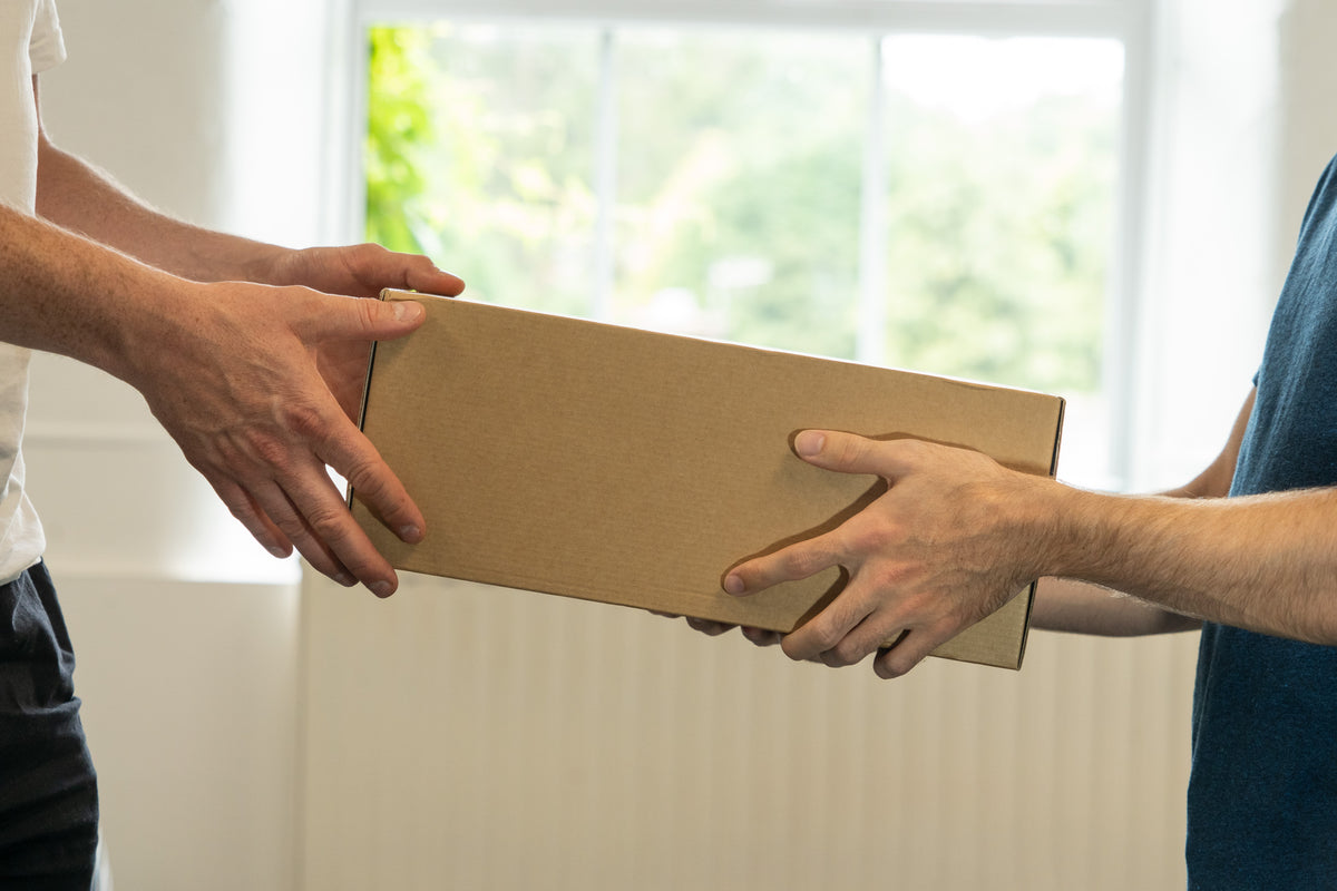A plain cardboard parcel passing from one person's hands to another's indoors, in front of a bright window