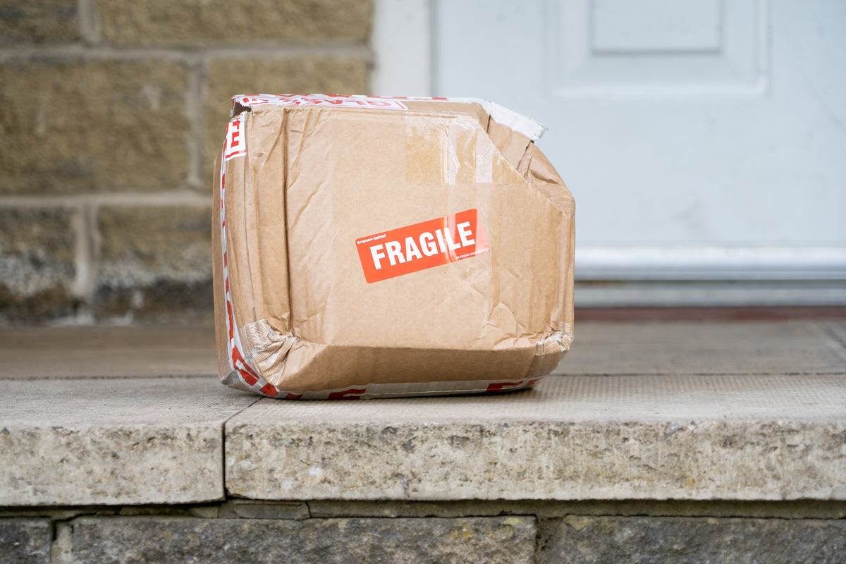 A crushed cardboard parcel with an orange FRAGILE label, left on a stone doorstep with one corner pushed inward
